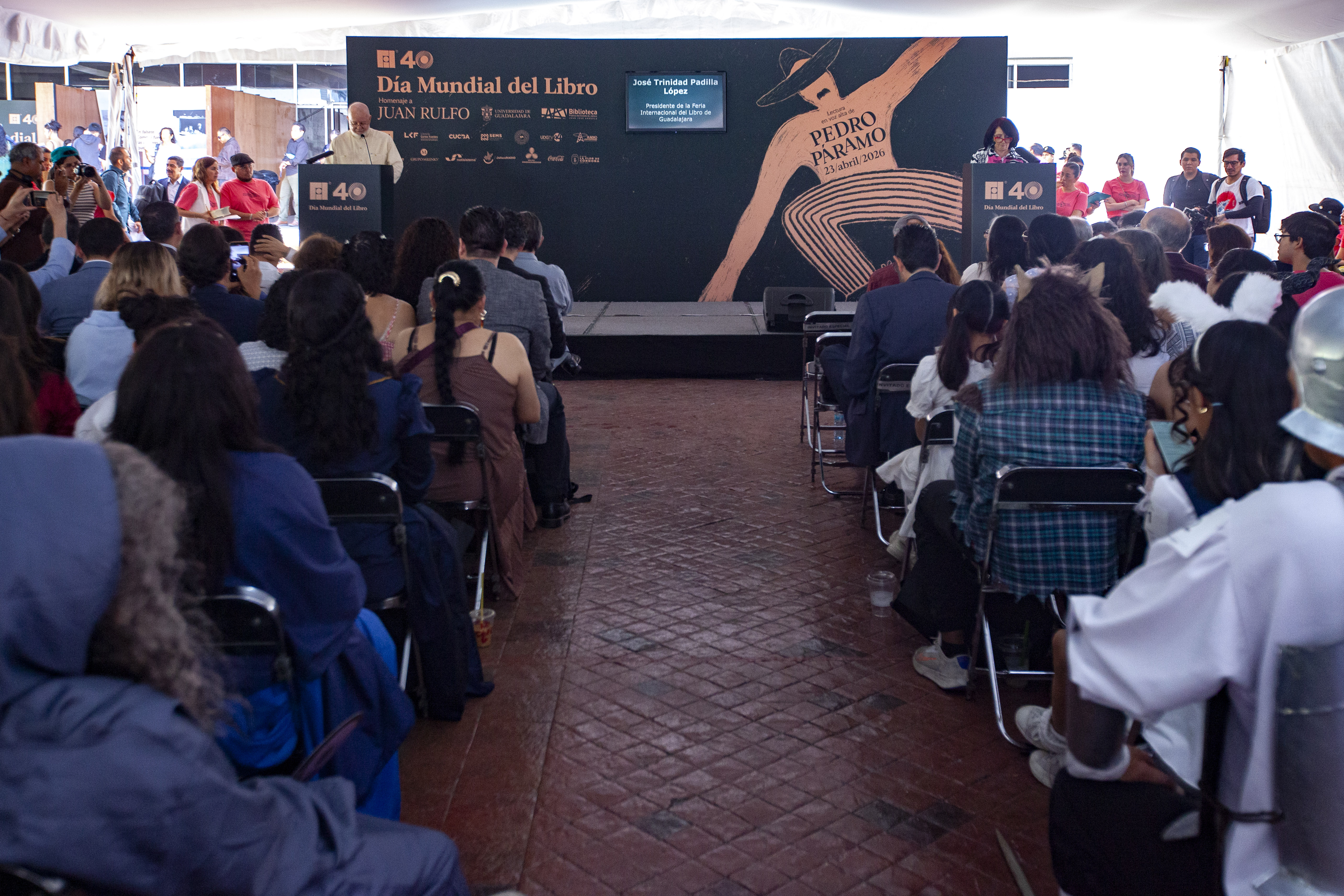 vista trasera asistentes a evento sentados autoridades del evento en escenario de fondo